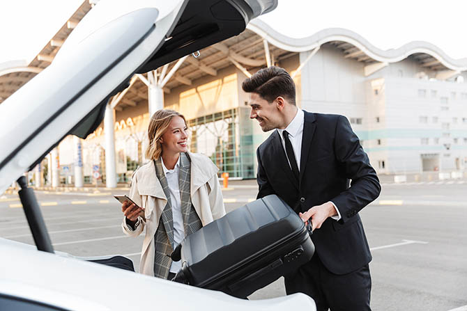 Image of young caucasian businesslike man and woman in formal wear putting travel luggage in car trunk outdoors